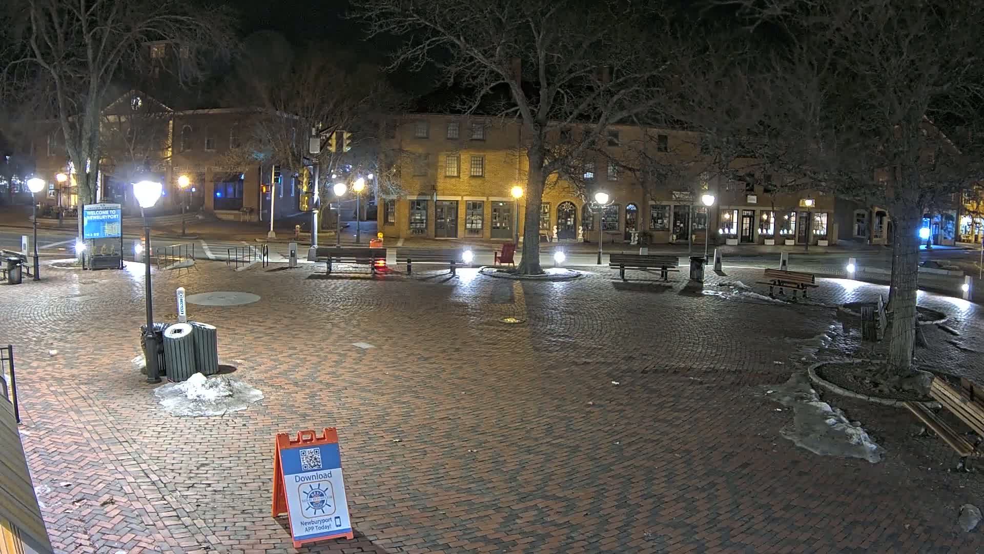 A brick town square at night, illuminated by streetlights, with patches of snow and ice on the ground, and several benches and trees surrounding it.