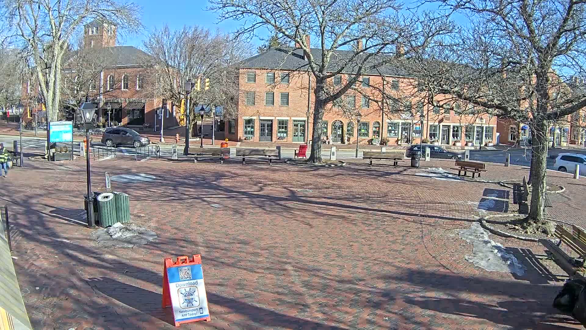 A brick town square with benches and trees, under a sunny winter sky with patches of snow and ice on the ground, is surrounded by brick buildings.