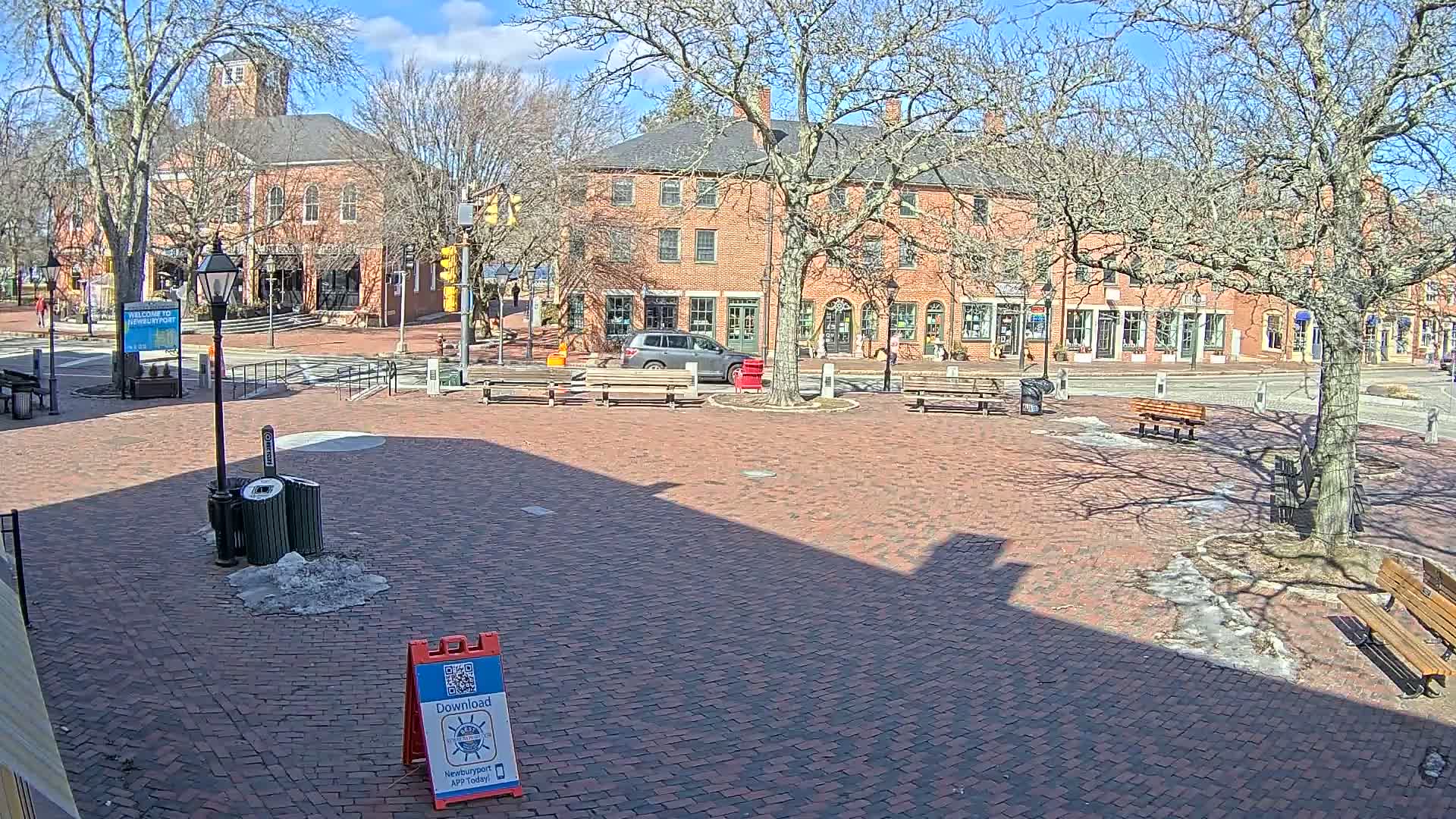 A brick town square with benches, trees, and a few cars parked near brick buildings on a sunny day with patches of snow and ice on the ground.