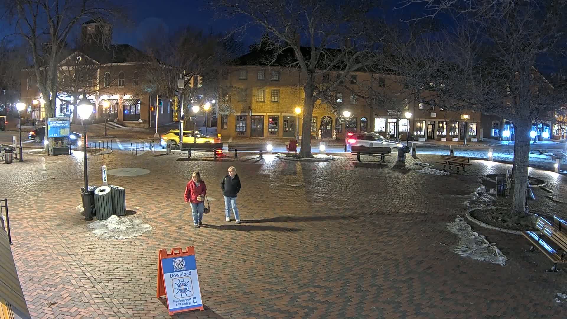 Two women walk across a brick plaza at night, under bare trees, past benches and buildings with some snow on the ground.