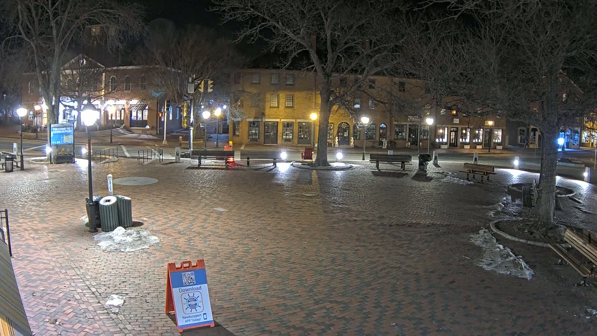 A brick town square at night, illuminated by streetlights, with patches of snow and ice on the ground, is surrounded by buildings and trees.