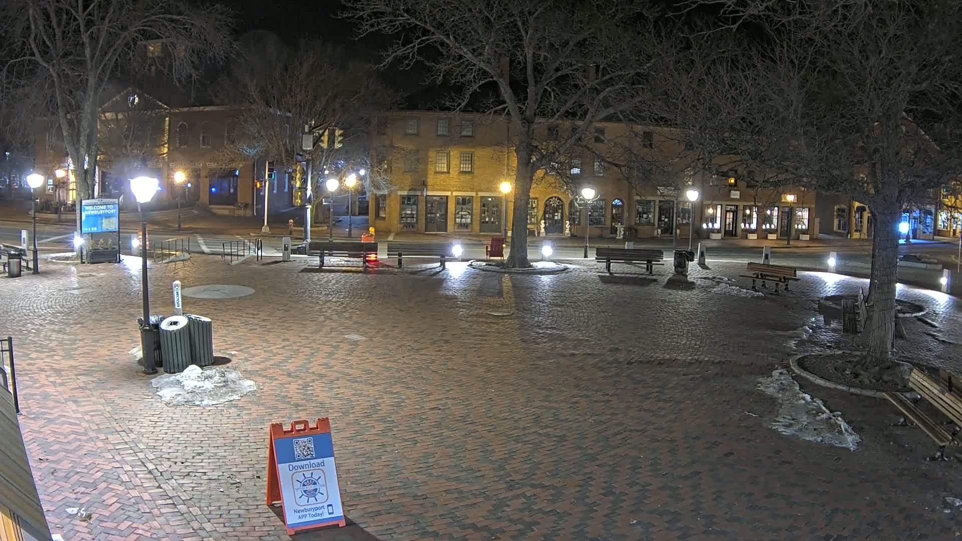 A brick plaza at night, illuminated by streetlights, with patches of snow and ice on the ground, is surrounded by buildings and bare trees.