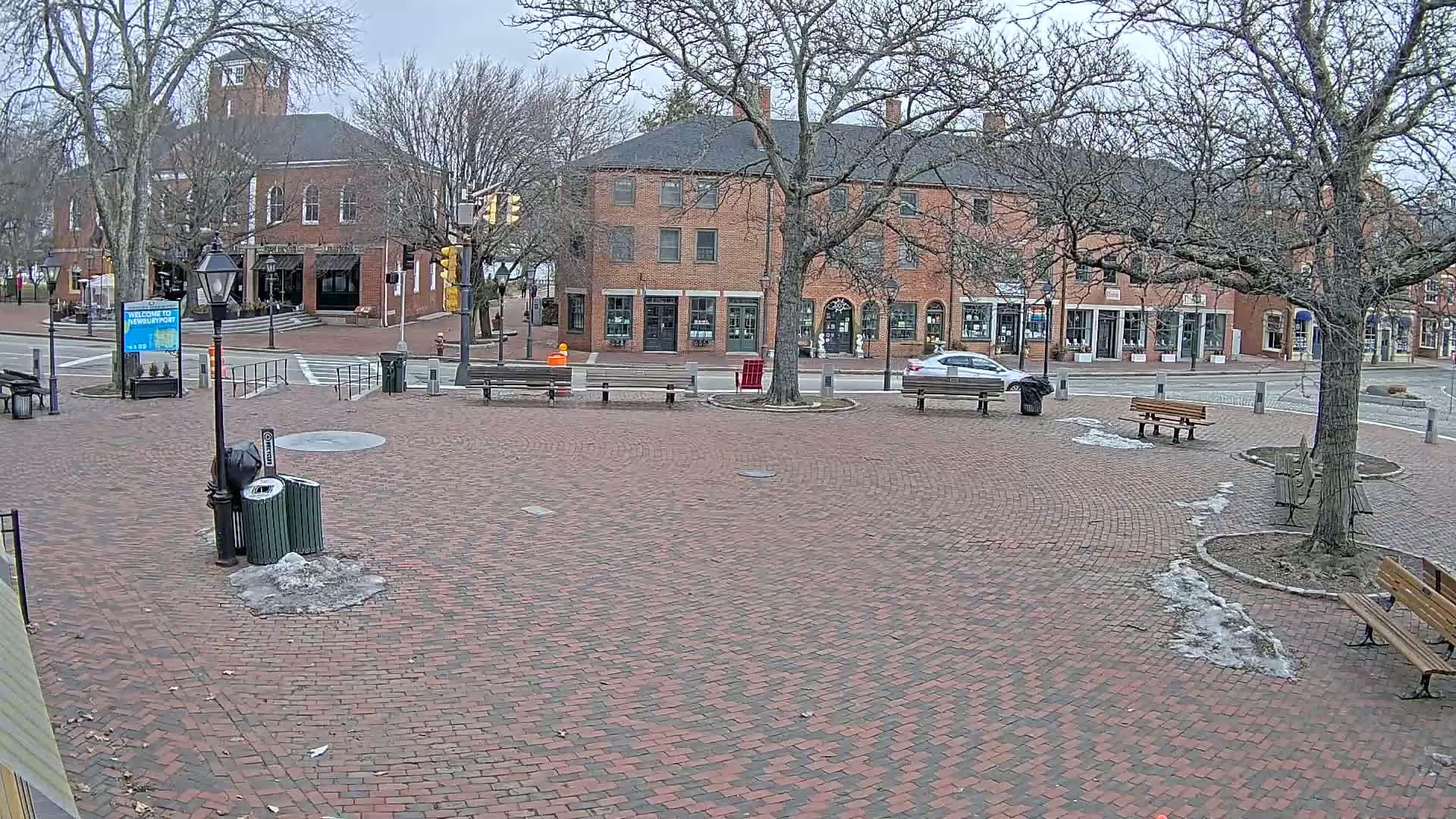 A brick town square with benches, trees, and a brick building under an overcast sky with patches of melting snow on the ground.