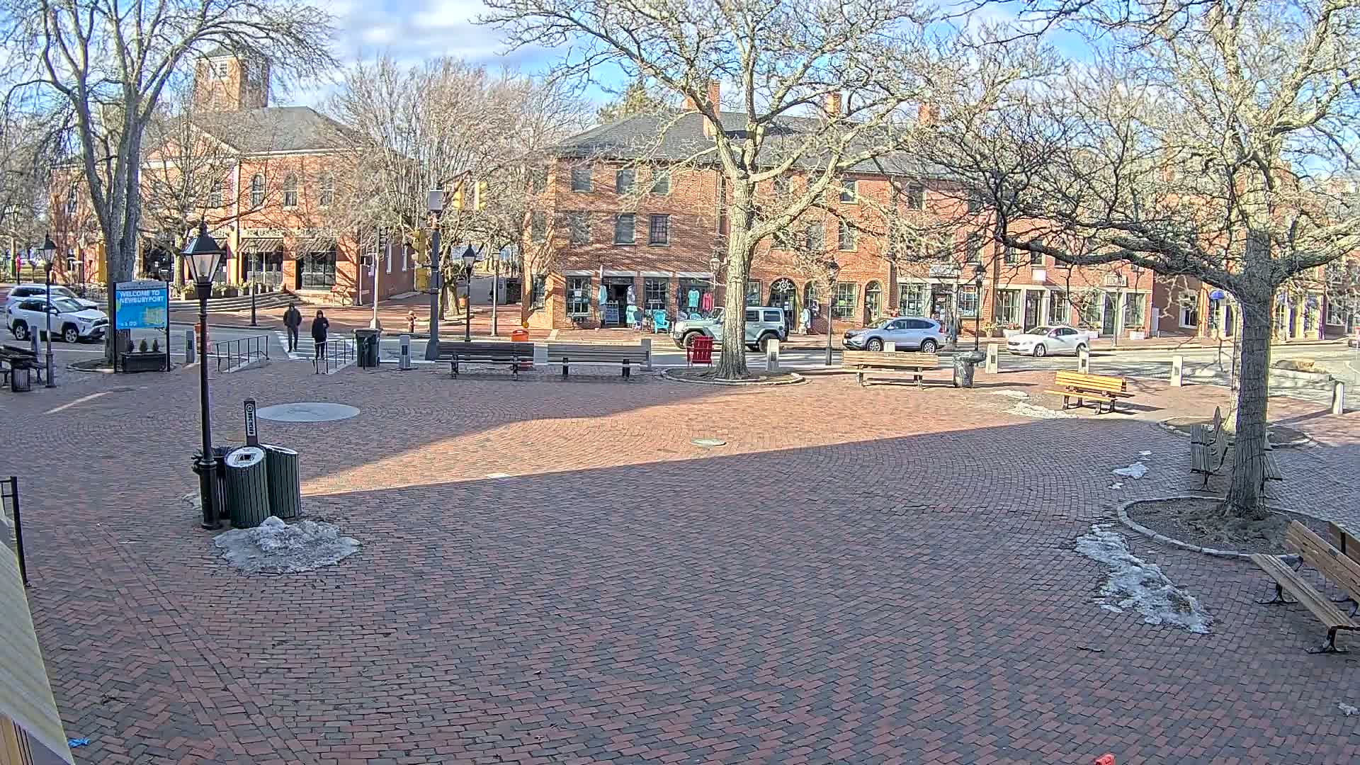 A brick town square with benches and bare trees under a sunny sky, shows some remaining patches of snow and ice on the ground.