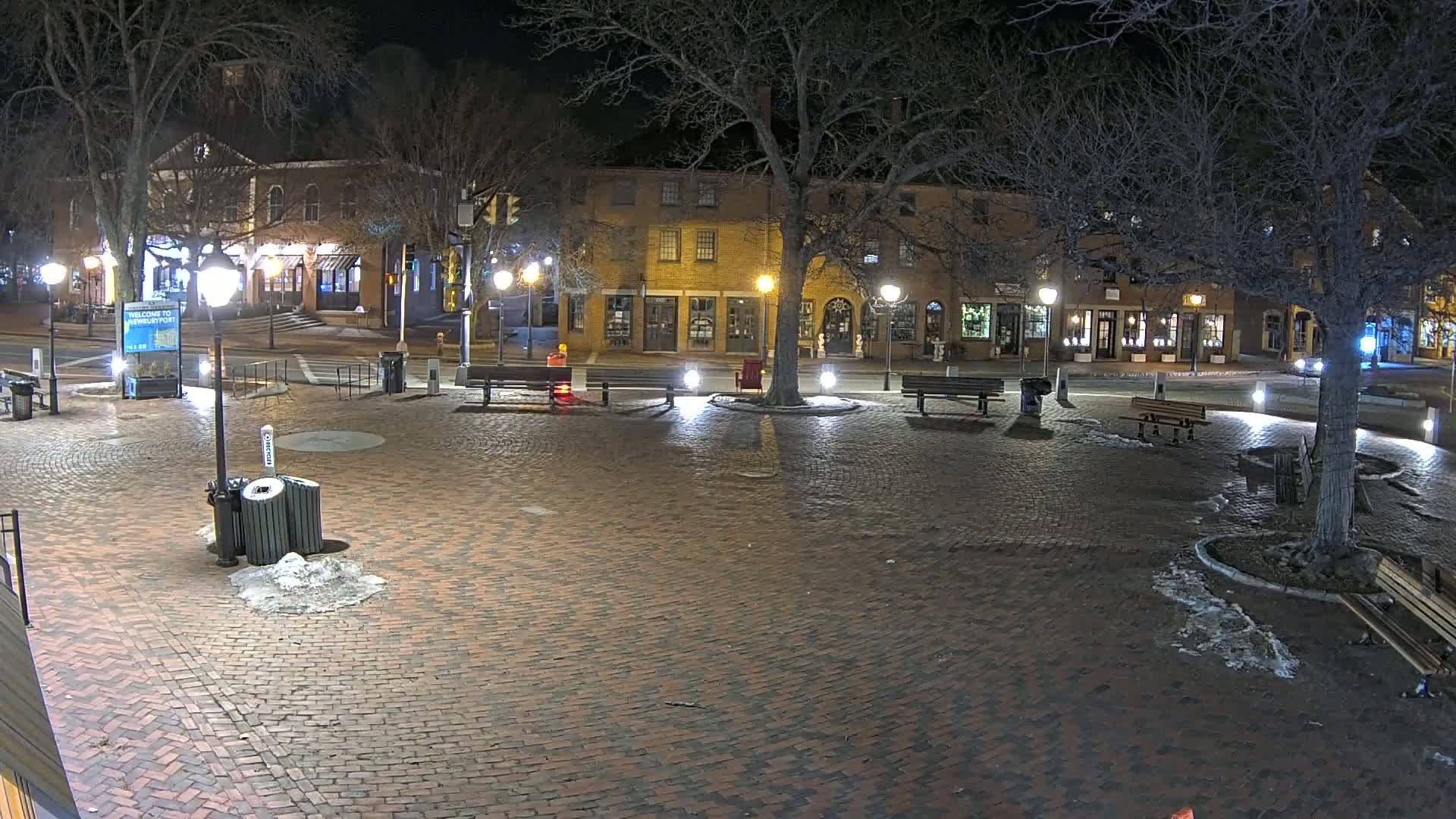 A brick town square at night, illuminated by streetlights, with benches, a tree, and some snow on the ground.