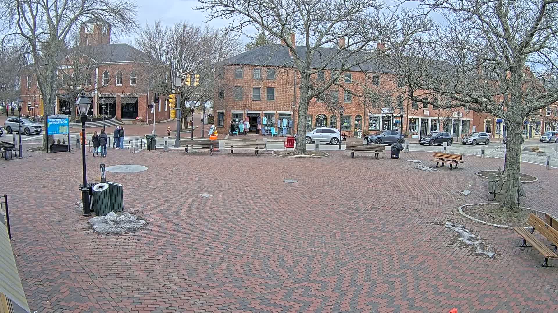 A brick town square with several parked cars and pedestrians, under a cloudy sky.