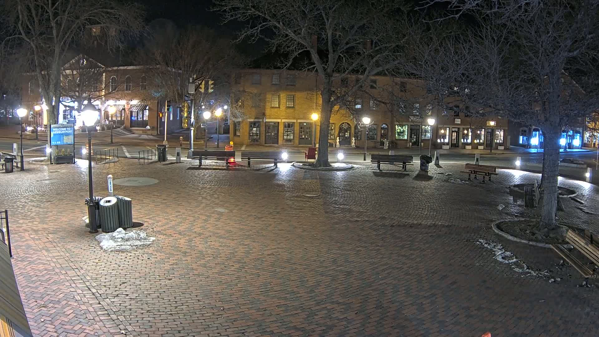 A brick plaza at night, illuminated by streetlights, features benches, a tree, and buildings lining the perimeter.