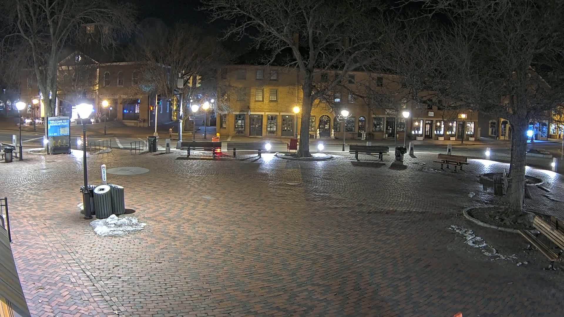 A brick plaza at night is illuminated by streetlights, with benches, trees, and a few parked cars visible in the background, and small patches of snow or ice on the ground.