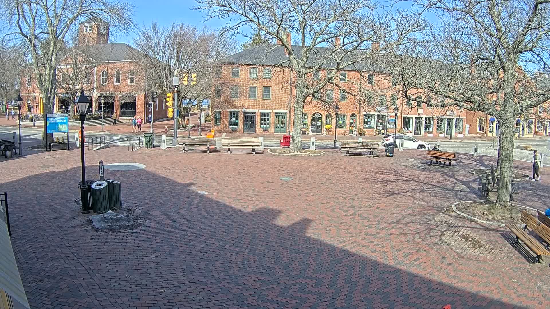 A brick town square with benches and trees, under a sunny sky, is surrounded by brick buildings.
