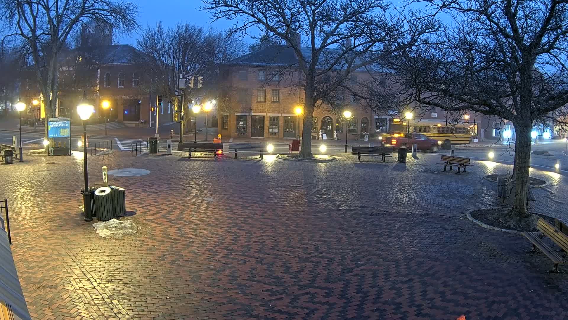 A brick town square at twilight is illuminated by streetlights, with benches, trees, and buildings visible in the background.