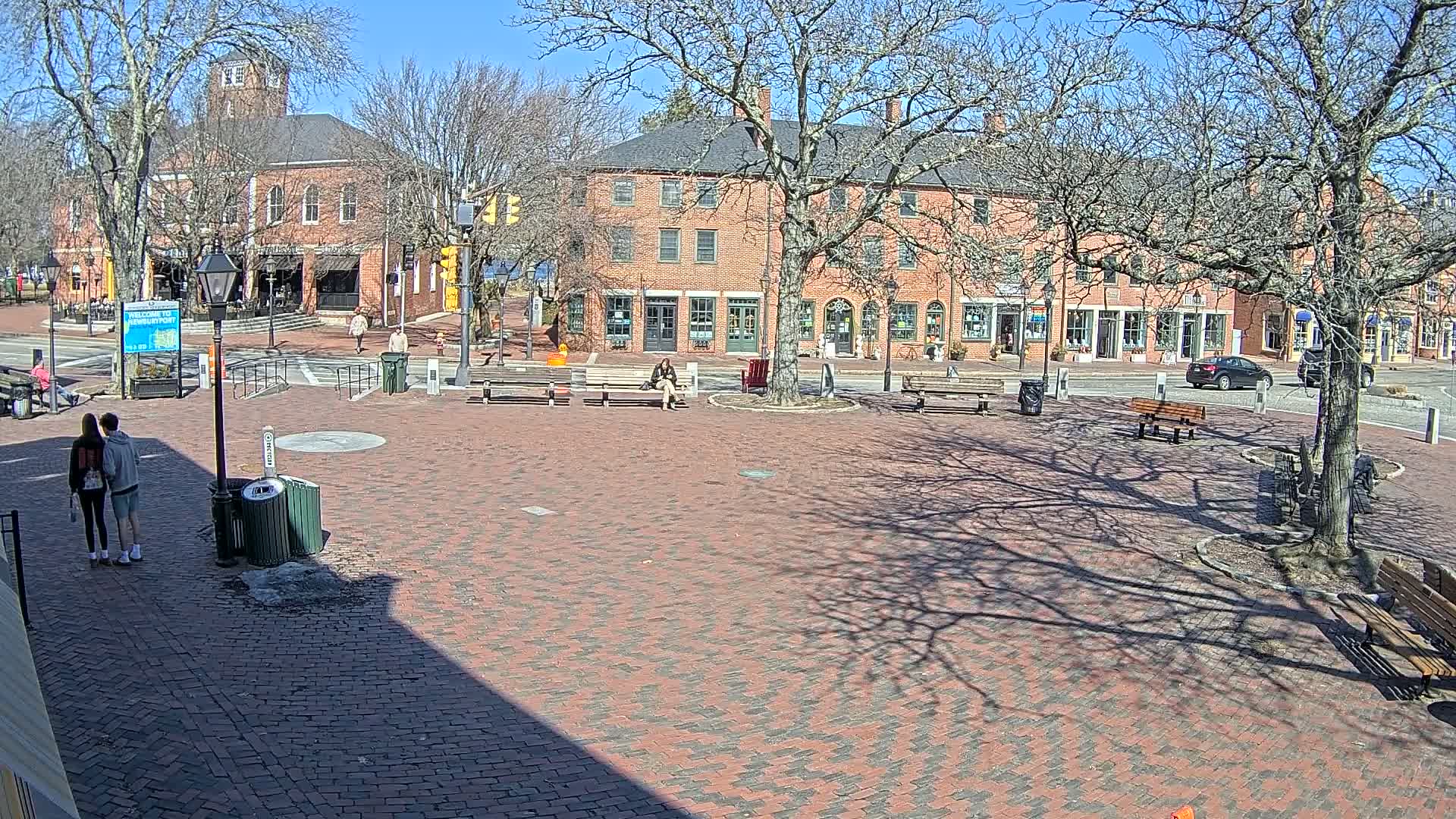 A brick town square with benches and several brick buildings under a sunny, clear sky.