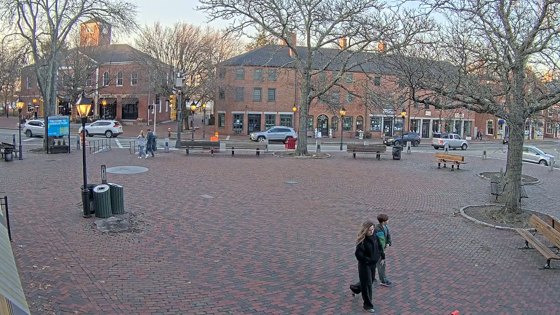 A brick plaza in a town square, under a partly cloudy evening sky, features several parked cars, benches, pedestrians, and a brick building with shops on the street behind it.