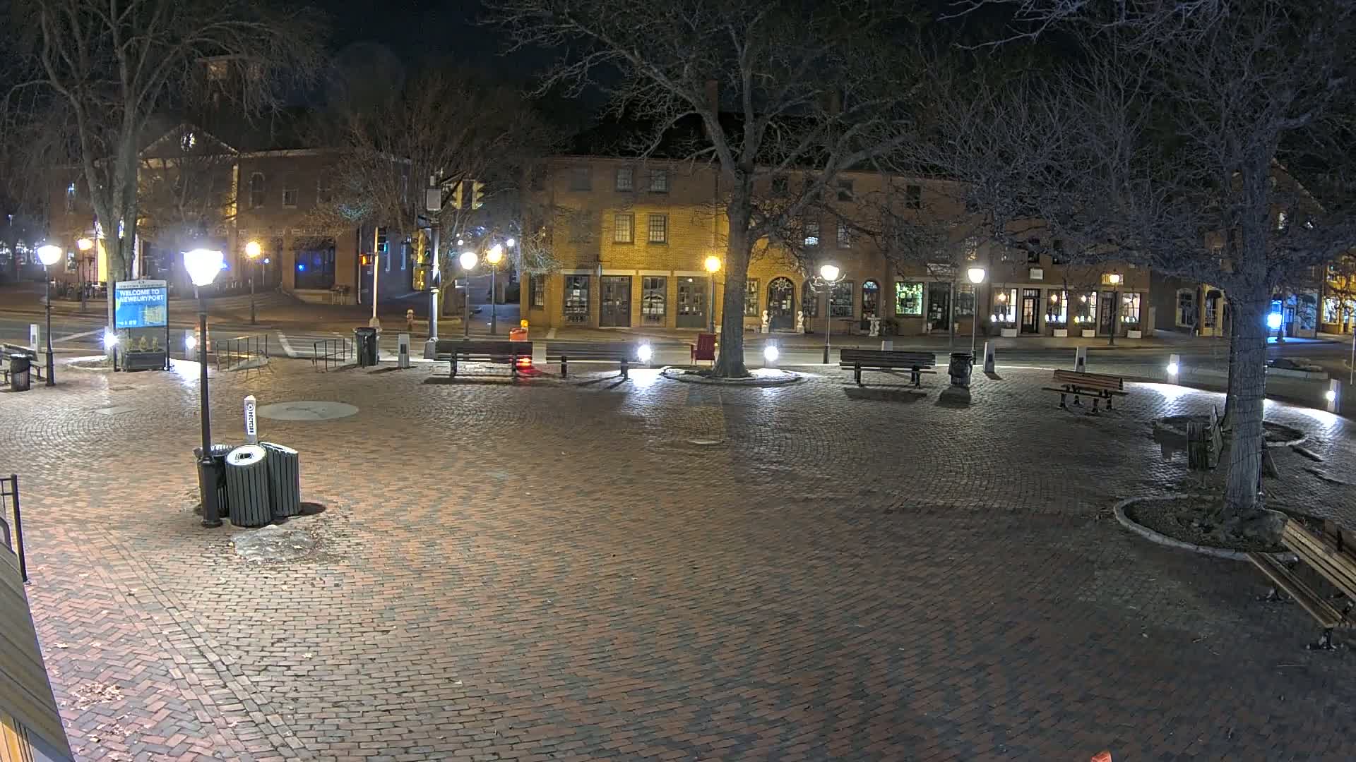 A brick plaza at night, illuminated by streetlights, is surrounded by low buildings and bare trees.