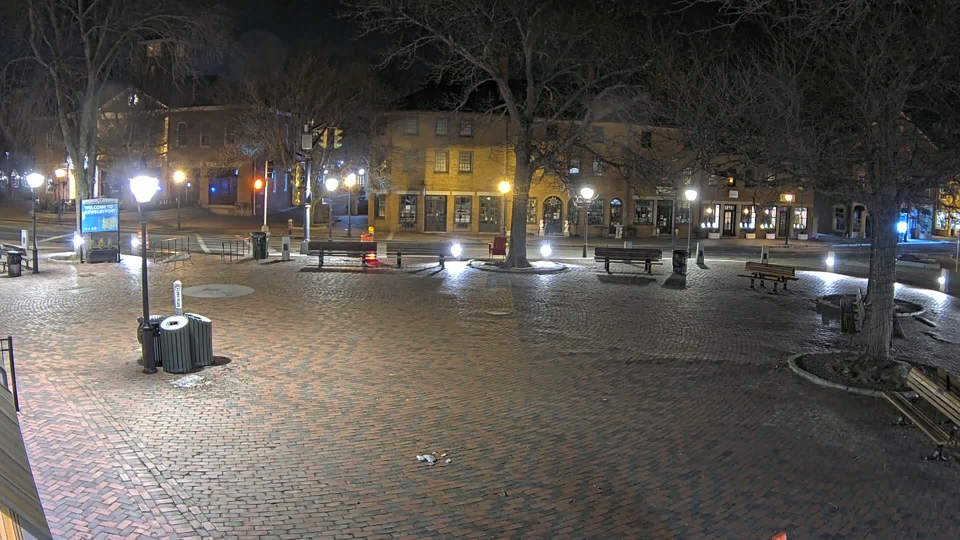 A brick plaza at night, illuminated by streetlights, is surrounded by buildings and trees, with several benches and trash receptacles present.