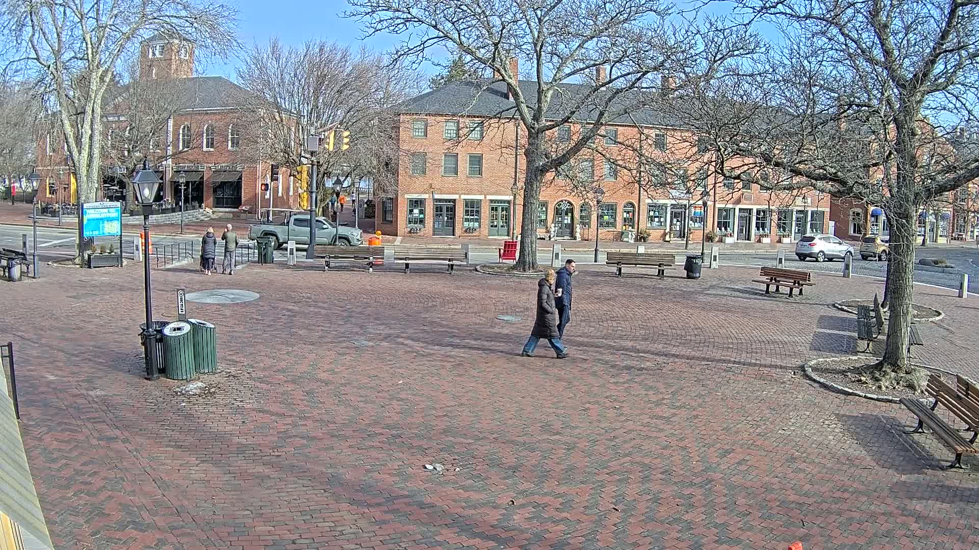 A brick town square with several people walking, benches, and trees, under a sunny winter sky.