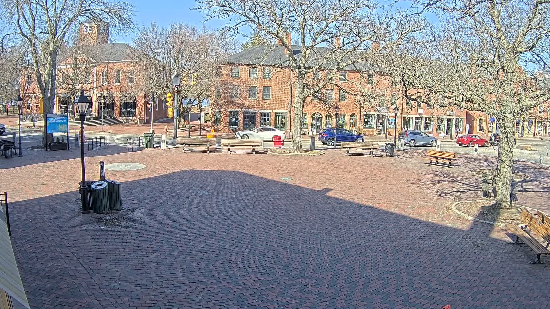 A brick plaza with several benches and a few parked cars is situated in front of brick buildings on a sunny day with bare trees.