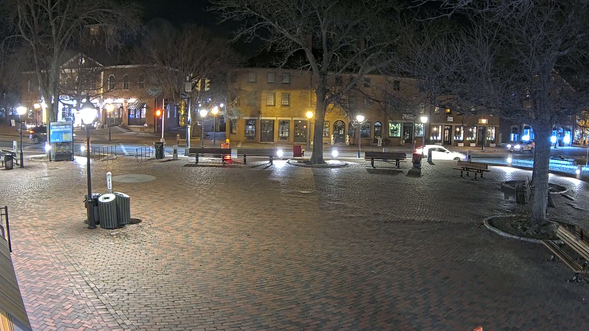A brick town square at night, illuminated by streetlights, with a few cars and empty benches surrounding a central tree, under a dark, clear sky.