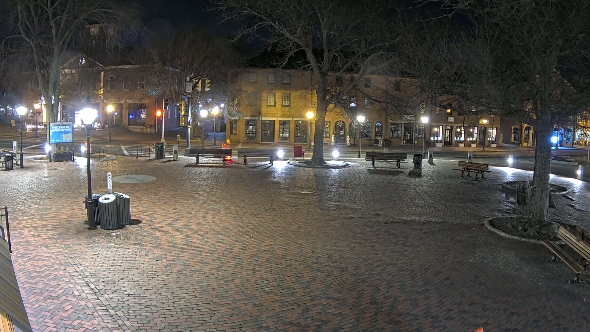 A brick plaza at night, illuminated by streetlights, is surrounded by buildings and trees, with several benches and trash cans visible.