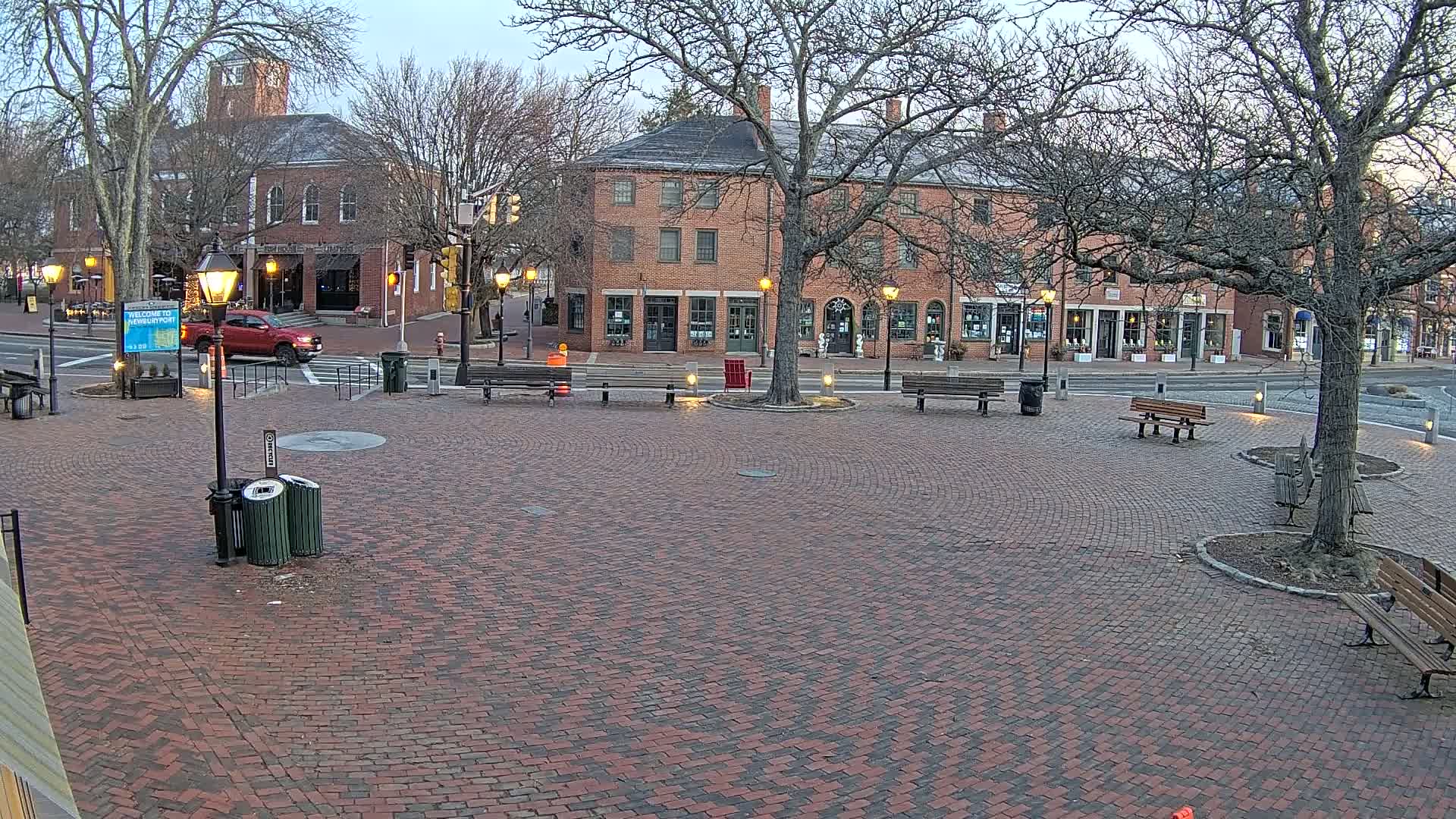 A brick plaza with benches and street lamps is situated in front of brick buildings under a mostly cloudy, dusky sky.