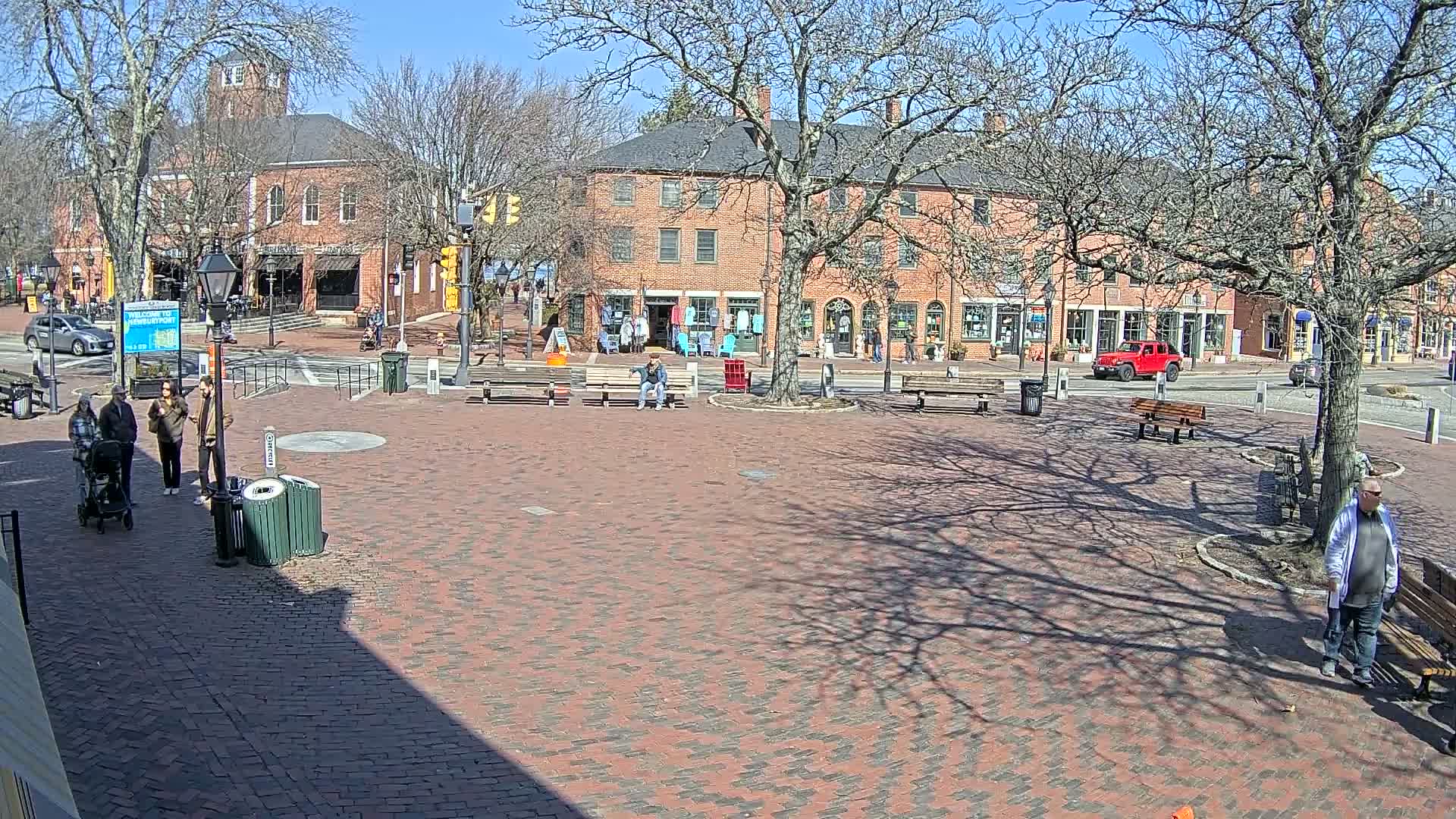 A brick town square with several people walking and sitting on benches, under a sunny sky with bare trees.
