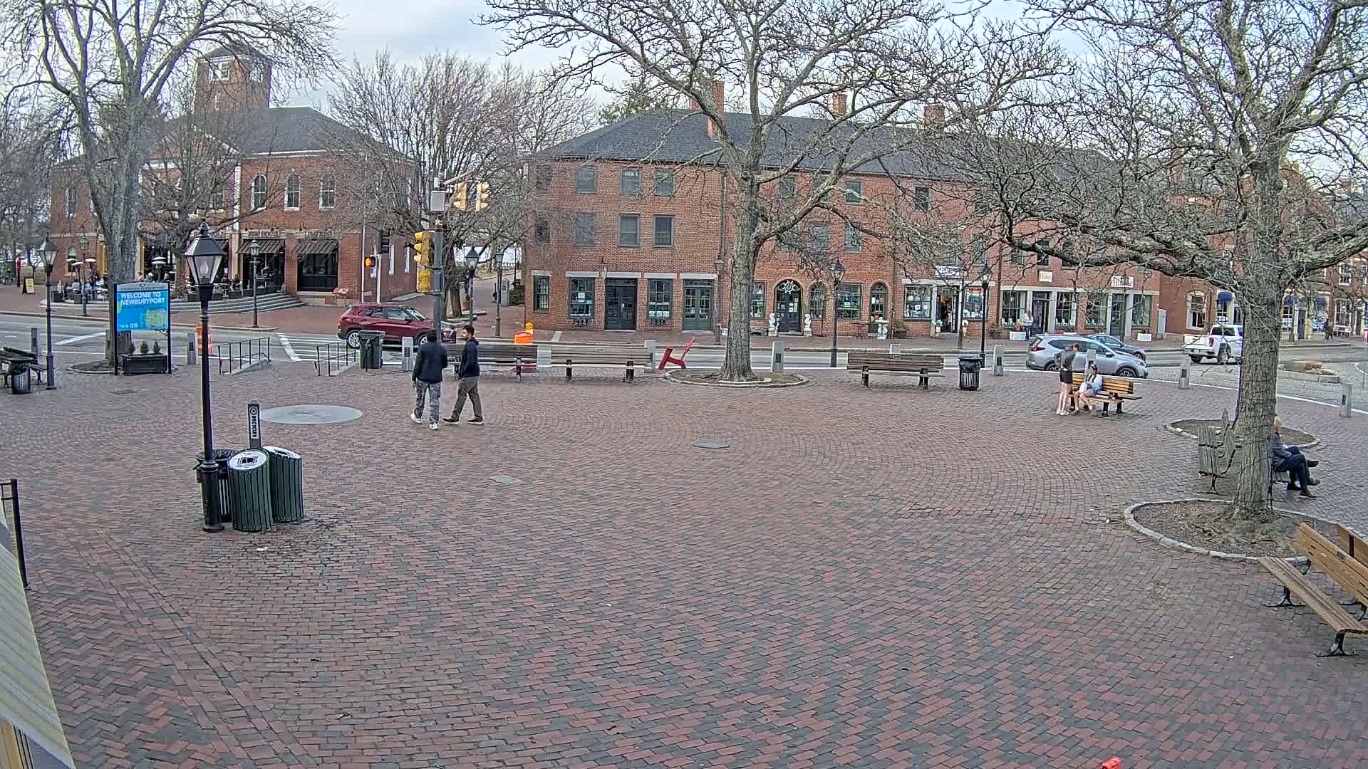 A brick town square with several people and cars visible on a partly cloudy day, surrounded by brick buildings and bare trees.