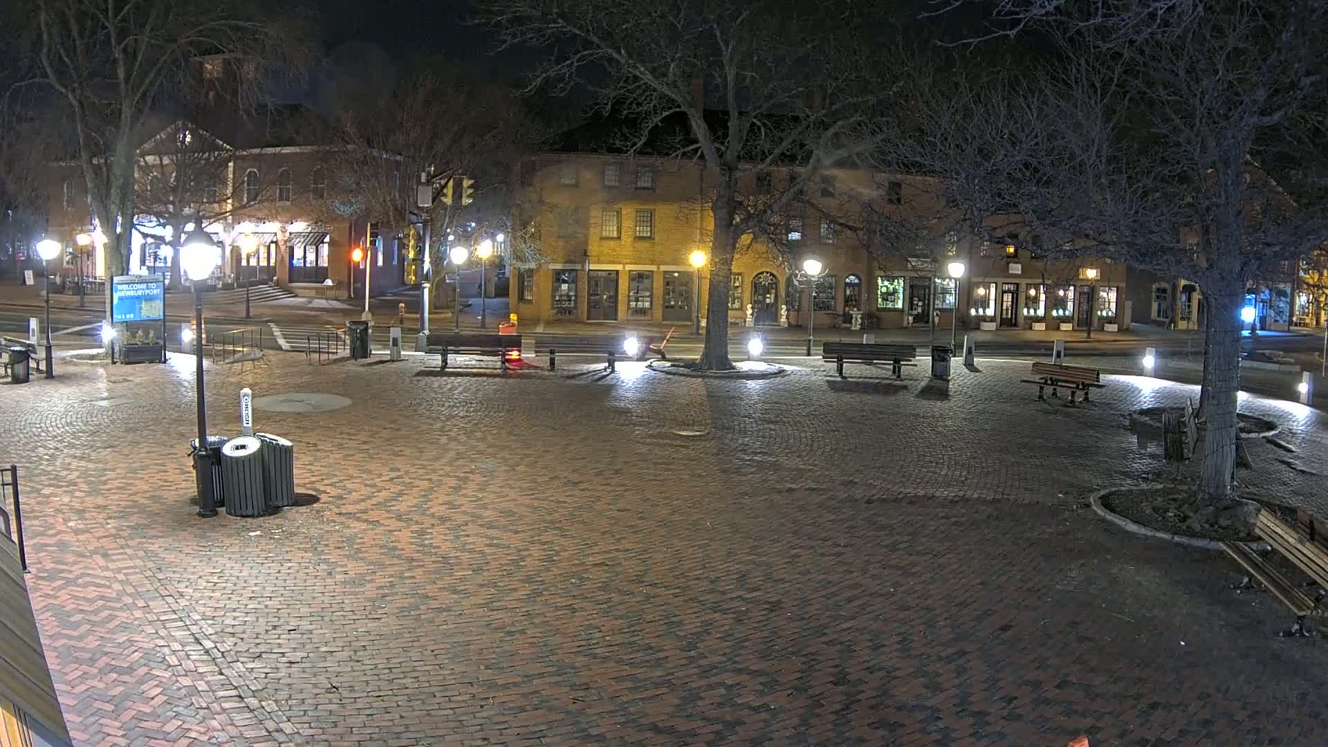 A brick town square at night, illuminated by streetlights, features several benches, a tree in the center, and buildings lining the perimeter.