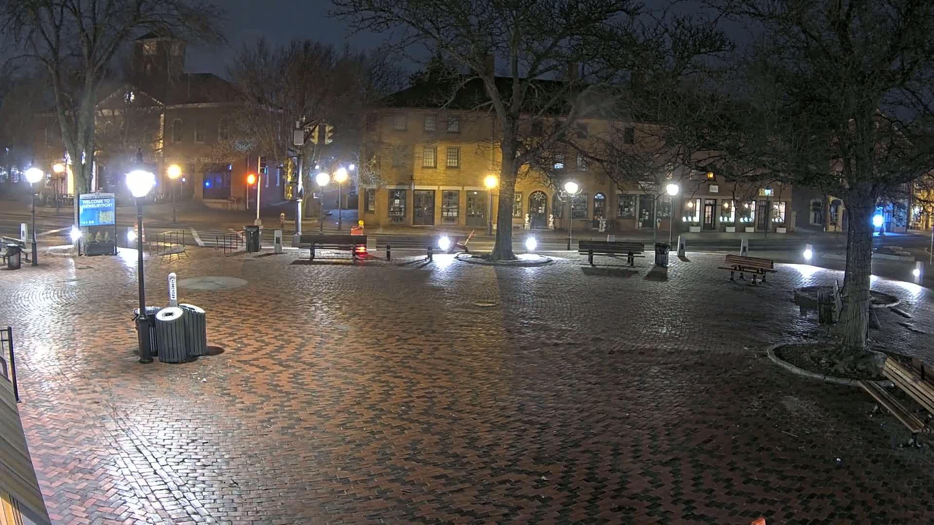 A brick plaza at night, illuminated by streetlights, with several benches and a tree in the center, and buildings lining the streets in the background.