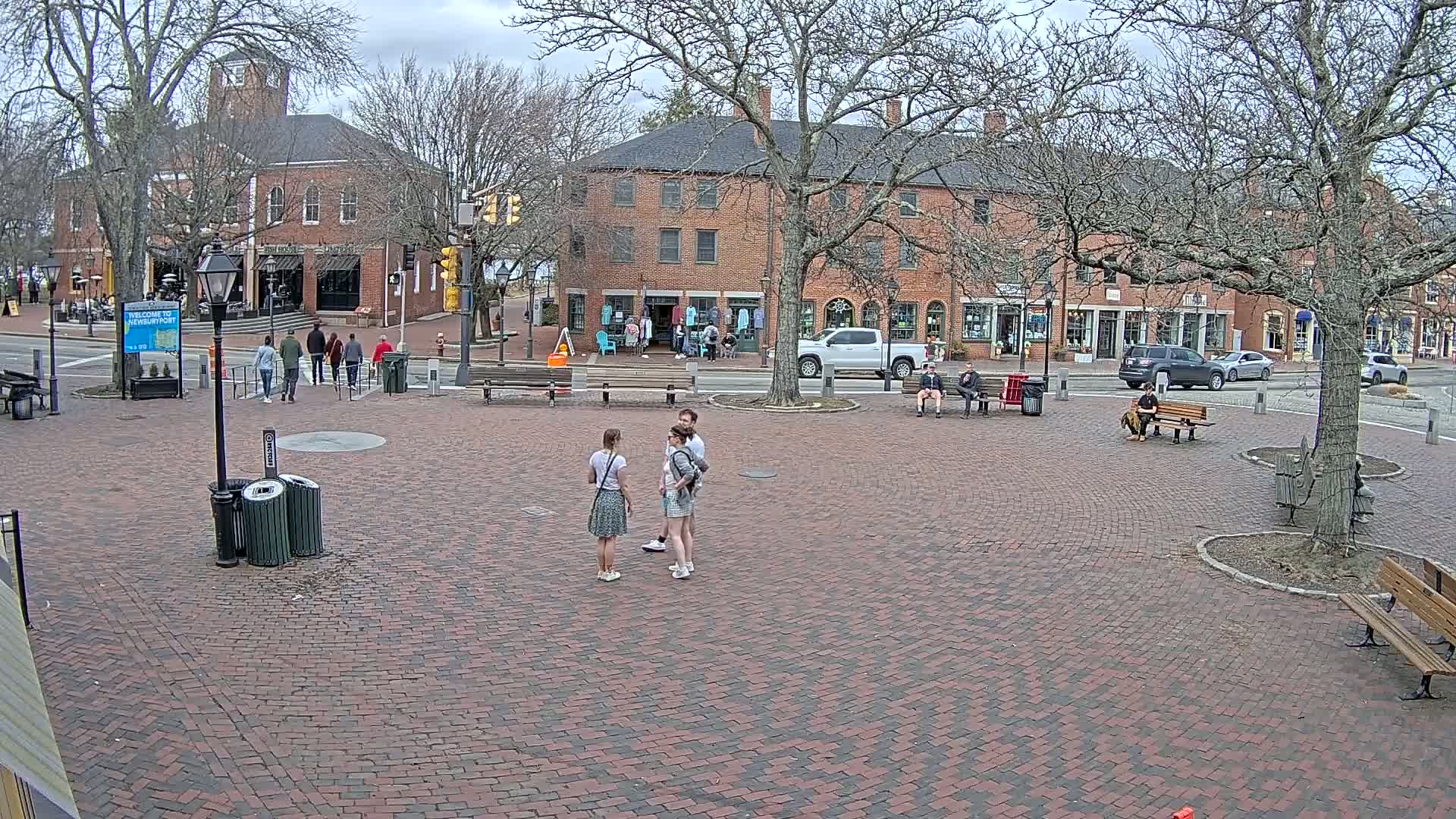 A brick town square with several people standing and sitting, under a mostly cloudy sky, surrounded by brick buildings and bare trees.