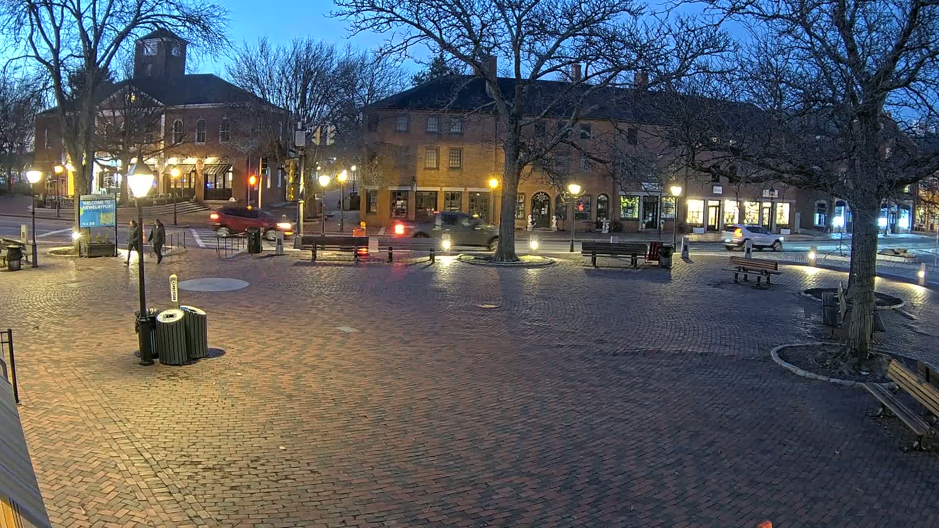 A brick town square at twilight, illuminated by streetlights, with several parked cars and pedestrians near brick buildings.