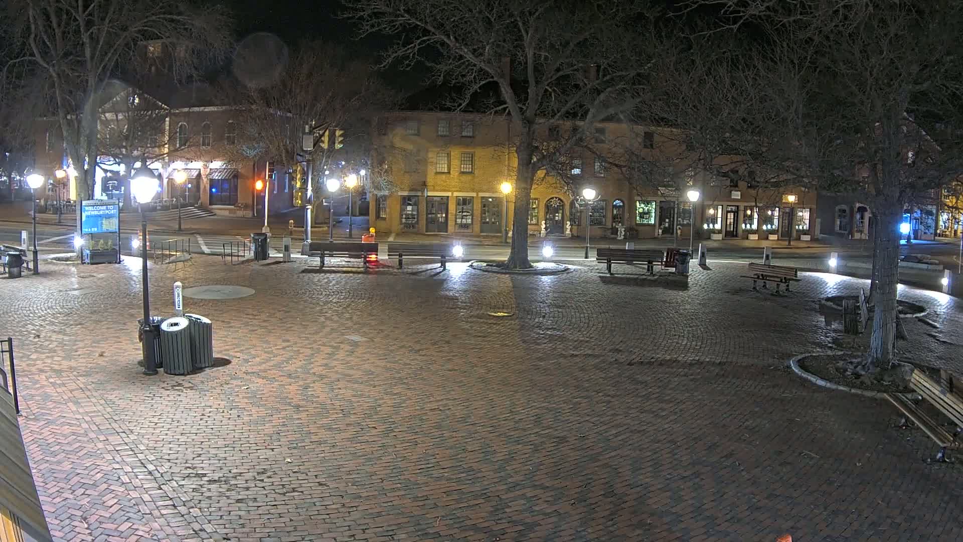 A brick plaza at night, illuminated by streetlights, is surrounded by buildings and trees, with a few benches and trash receptacles present.
