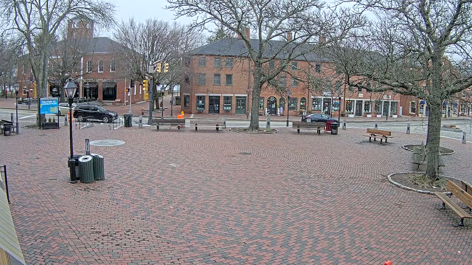 A brick plaza with several benches is surrounded by brick buildings under a cloudy sky.