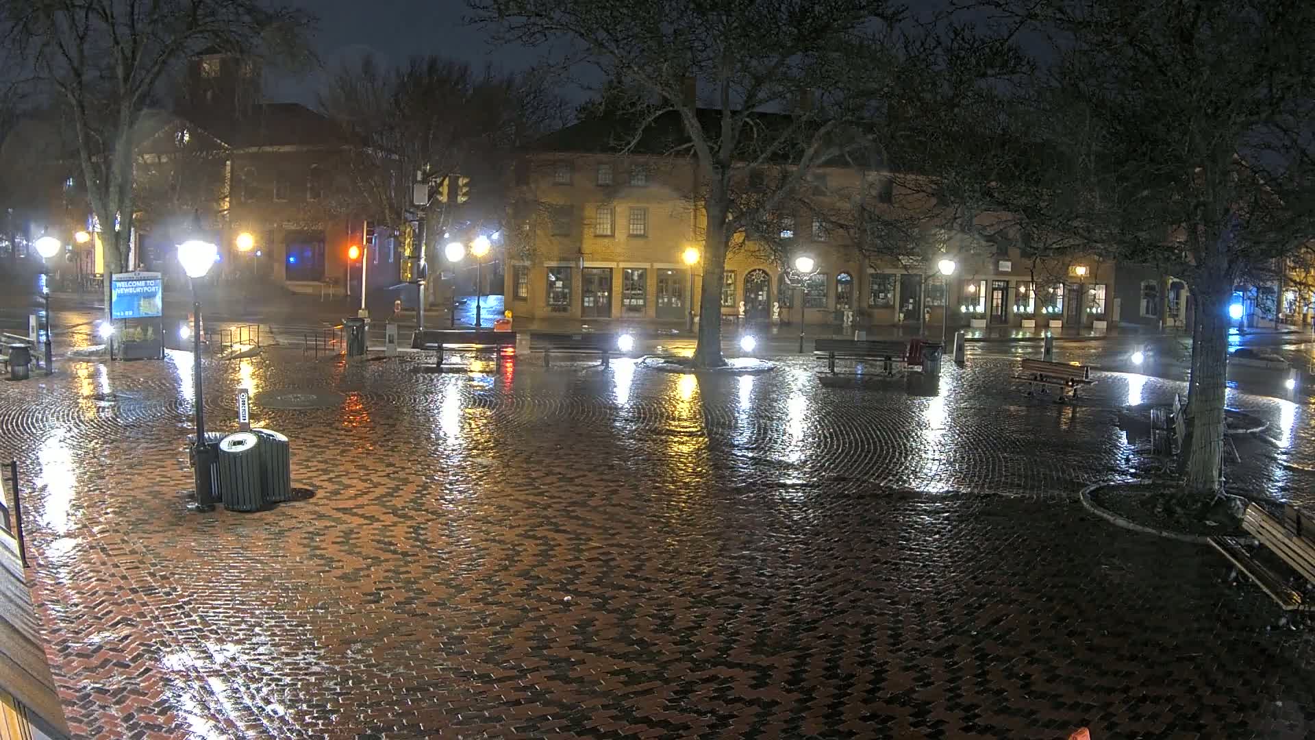 A rain-slicked brick town square at night is illuminated by streetlights reflecting on the wet cobblestones, with several benches and a tree in the center, and buildings lining the square's edge.