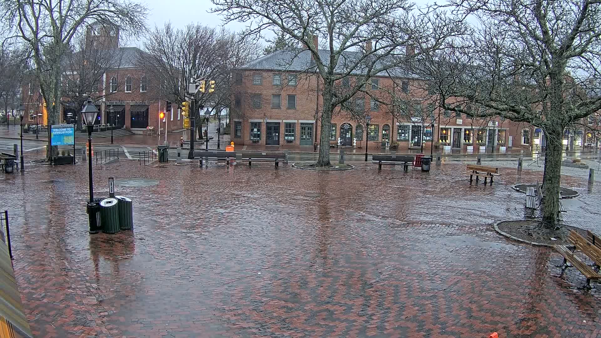 A brick town square, wet from rain, is surrounded by brick buildings and bare trees.