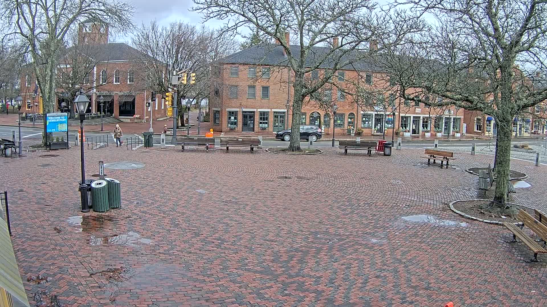 A brick plaza with benches and trees, situated in front of brick buildings on an overcast day with wet pavement.