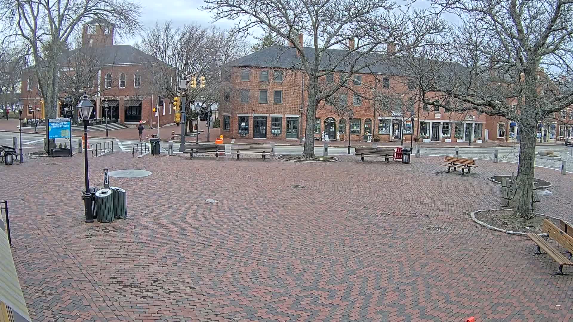 A brick plaza with several benches and trash cans, surrounded by brick buildings under a cloudy sky.