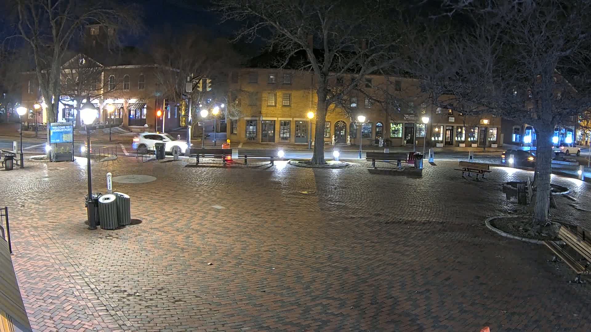 A brick town square at night, illuminated by streetlights, with a few cars and benches visible, under a clear, dark sky.
