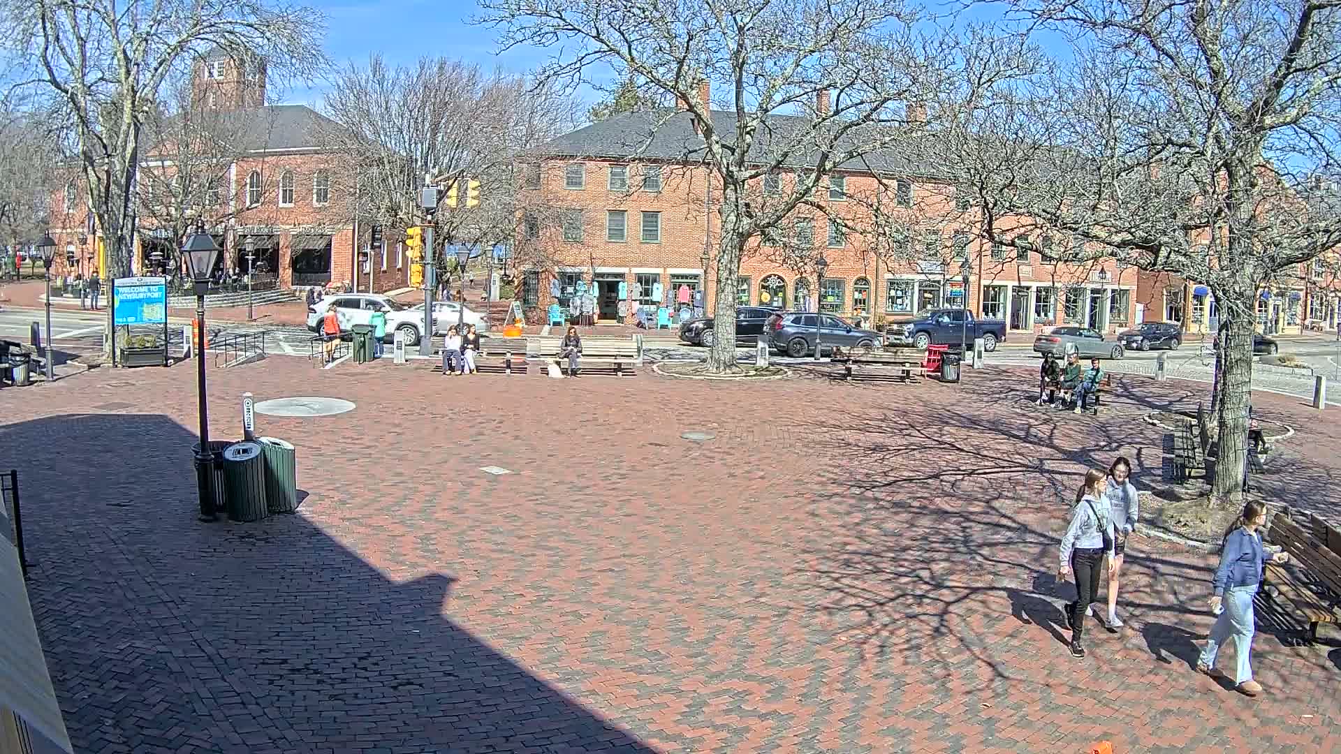 A brick town square with several people sitting and walking, surrounded by brick buildings and bare trees under a sunny sky.