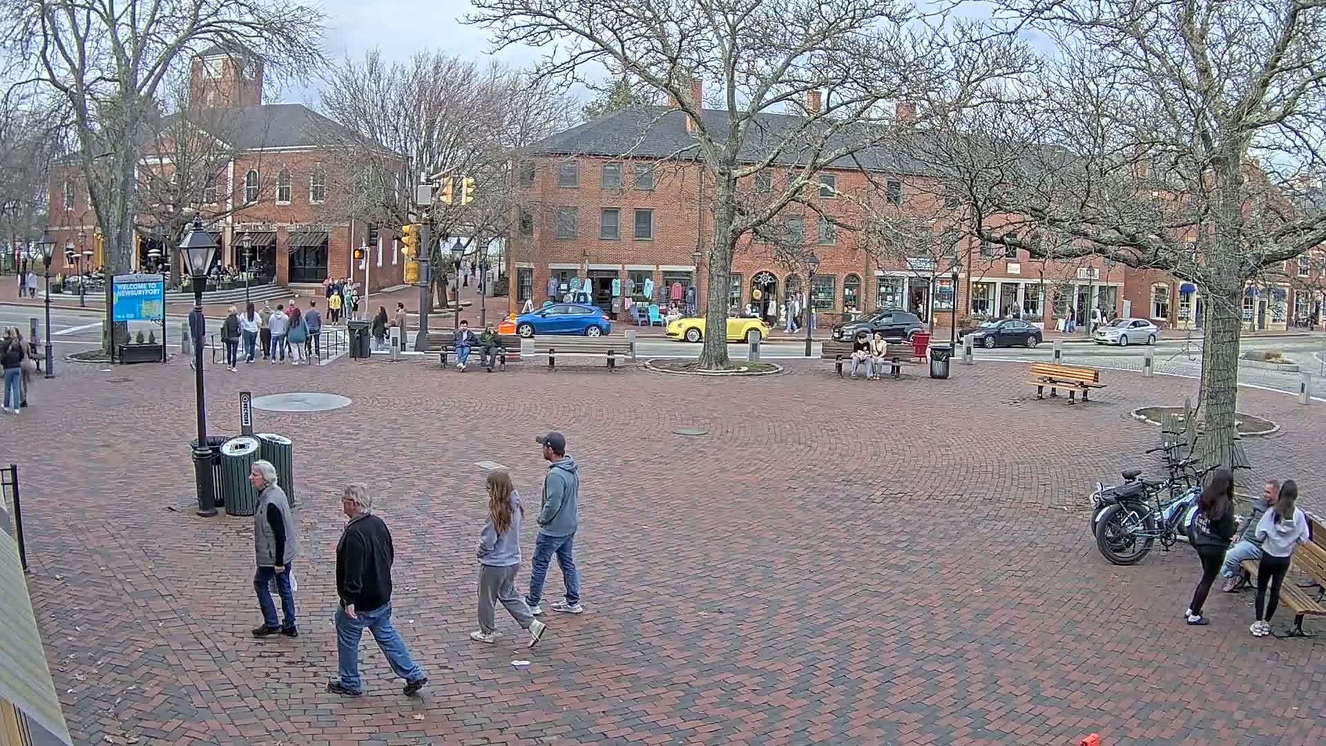 A brick town square with several people walking and sitting on benches, under a cloudy sky, surrounded by brick buildings and bare trees.