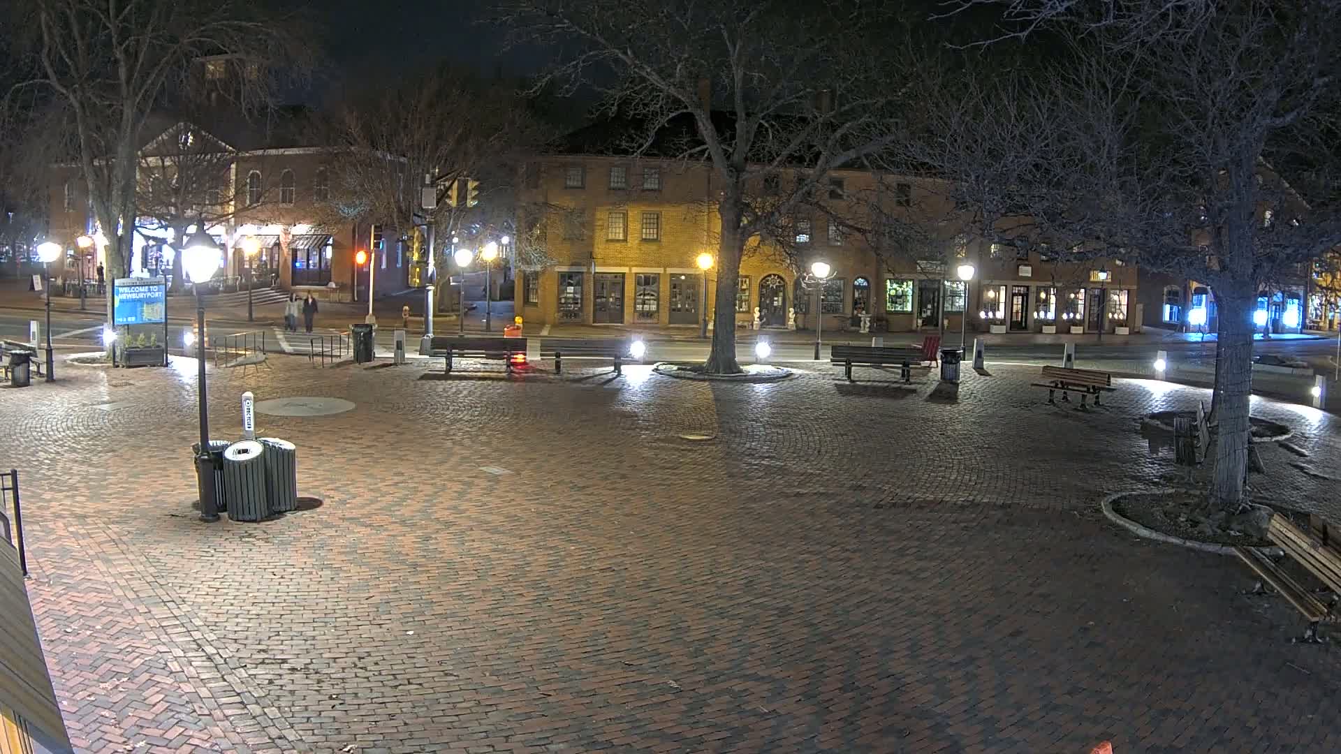 A brick plaza at night, illuminated by streetlights, with benches and trees, is surrounded by buildings.