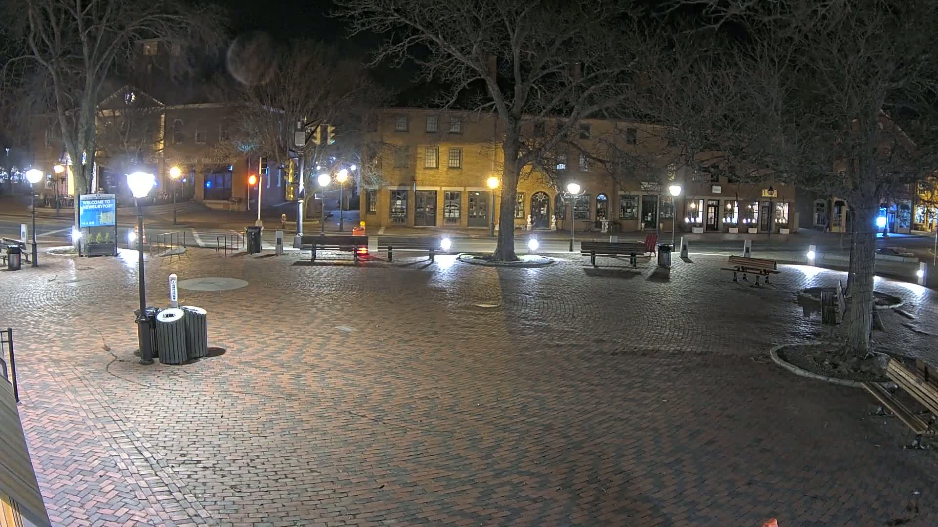 A brick plaza at night, illuminated by streetlights, is surrounded by buildings and trees under a clear, dark sky.
