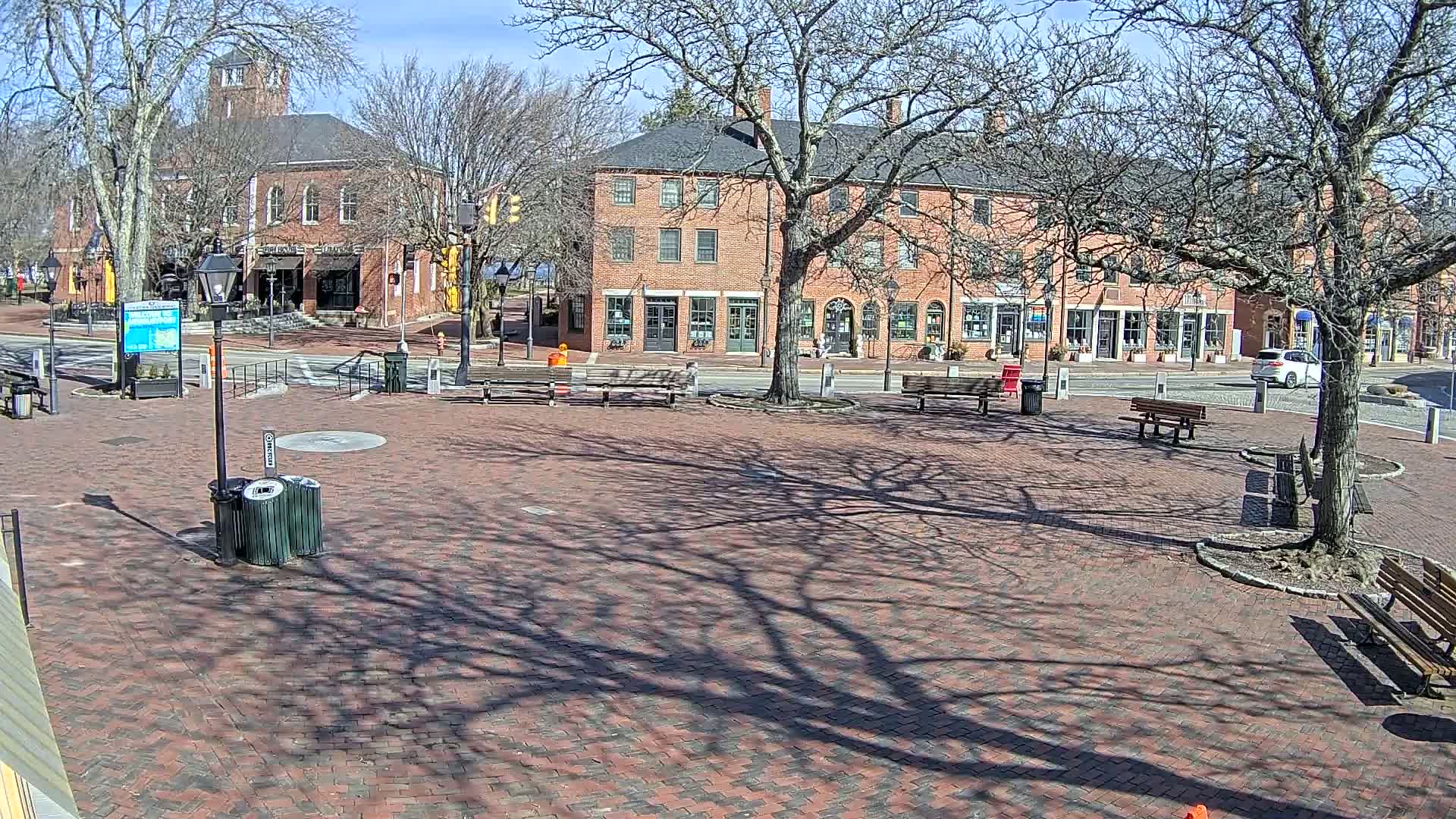 A brick town square with benches and trees casting shadows on a sunny day, featuring several brick buildings across a street.