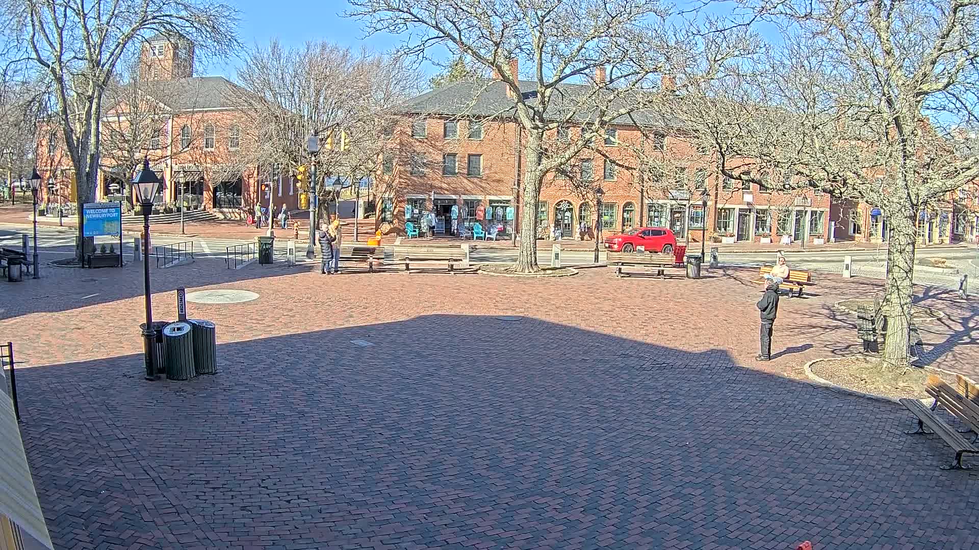 A brick town square with several people, benches, and brick buildings on a sunny day.