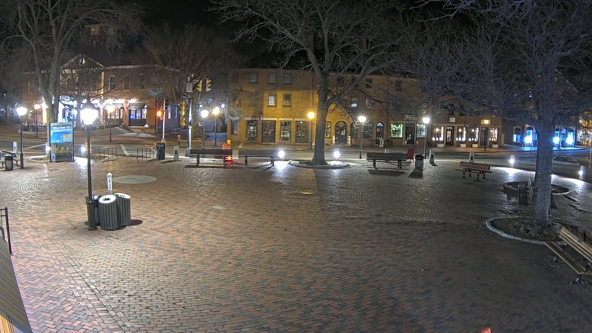 A brick plaza at night, illuminated by streetlights, with benches and trees surrounding a central area, and buildings visible in the background.