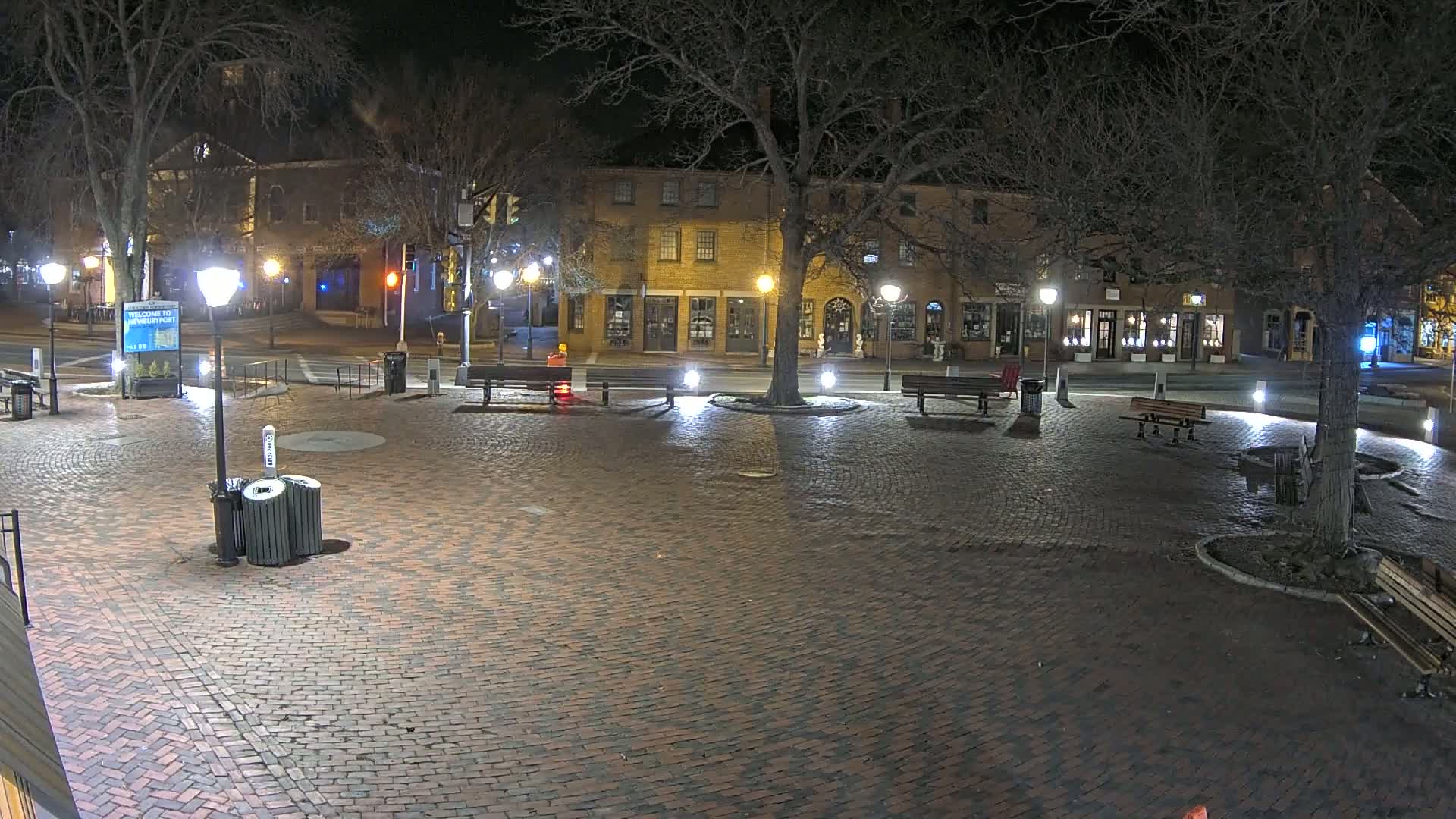 A brick plaza at night, illuminated by lampposts, is surrounded by buildings and trees.