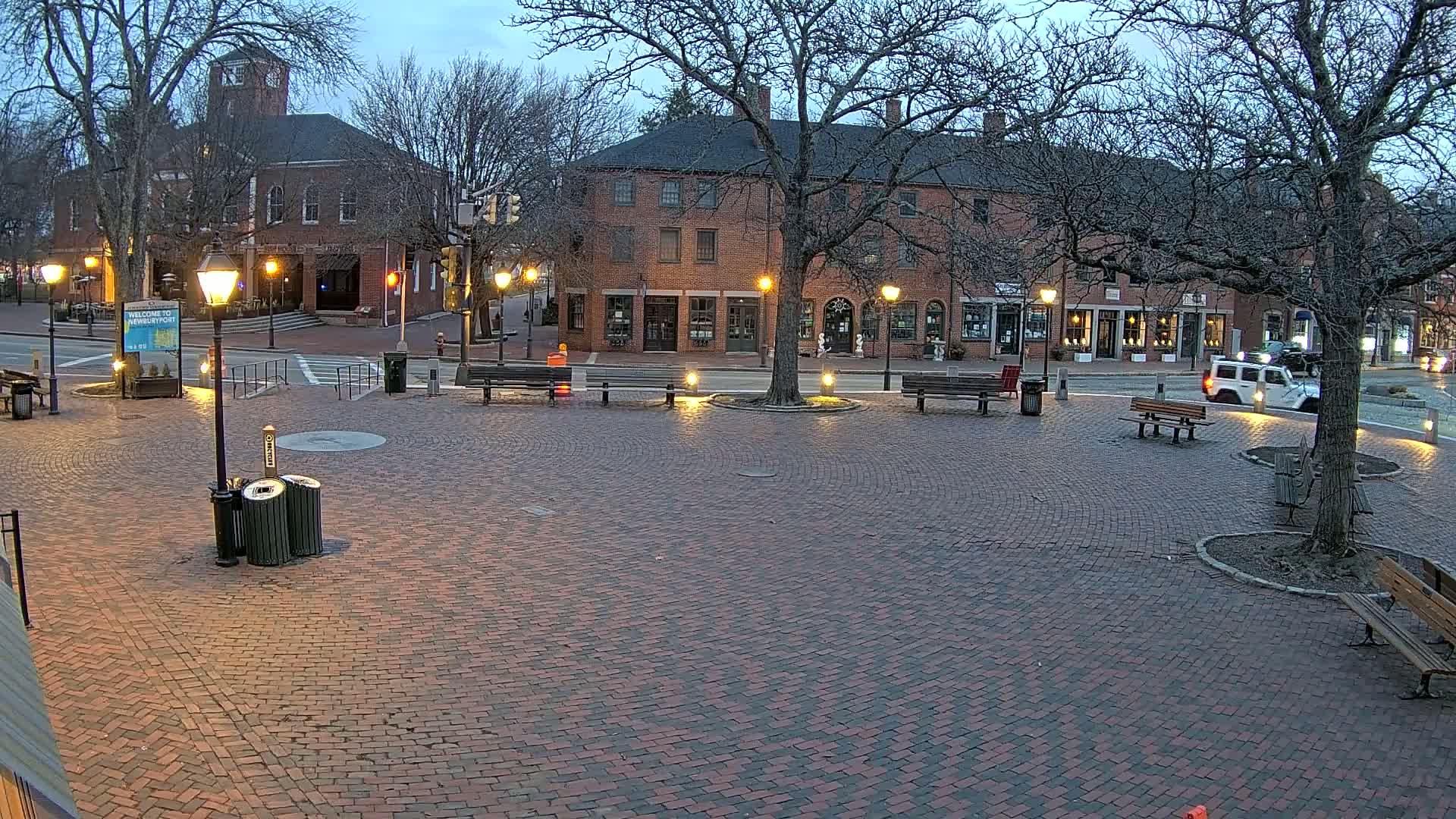 A brick plaza at twilight is surrounded by brick buildings and bare trees with several park benches and lit lampposts.