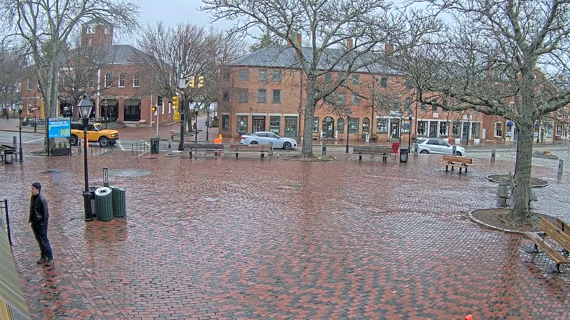 A brick town square, wet from rain, is surrounded by brick buildings and several parked cars, with a few people visible.