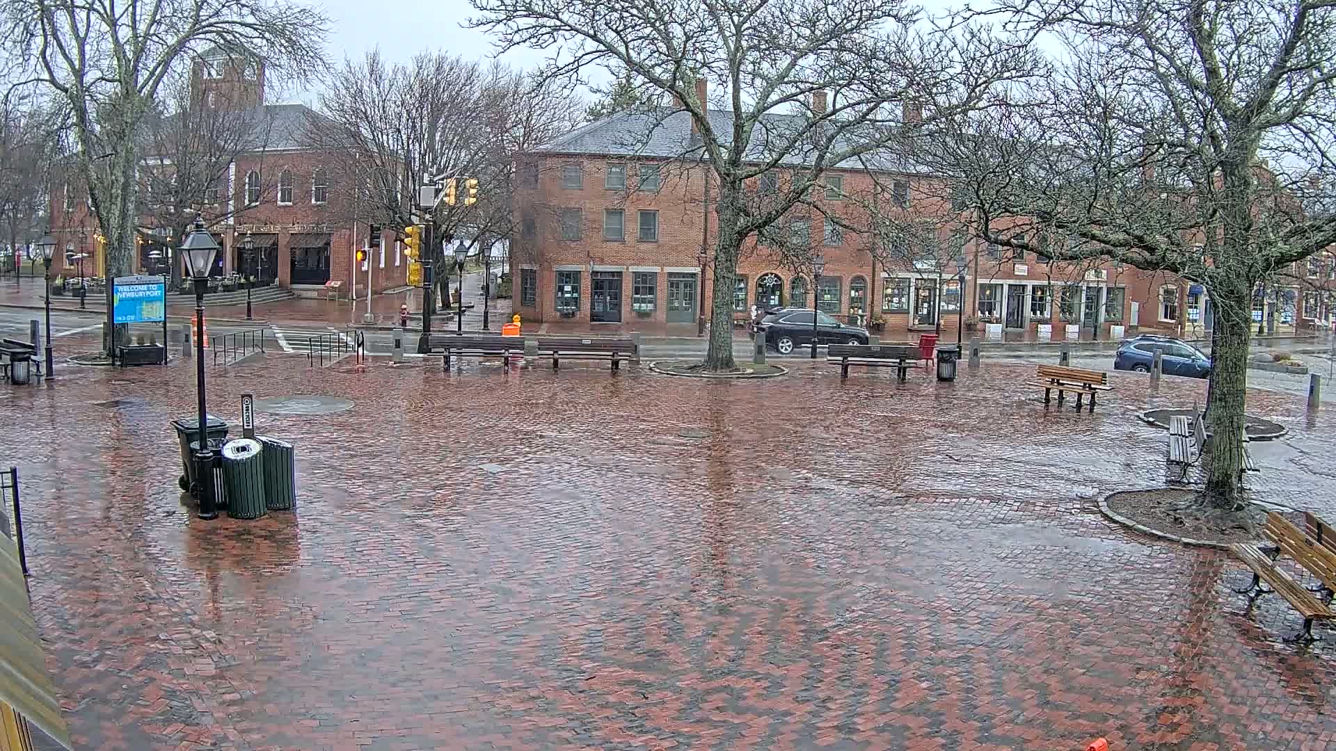 A brick town square is wet from rain, with several benches and buildings lining the perimeter.