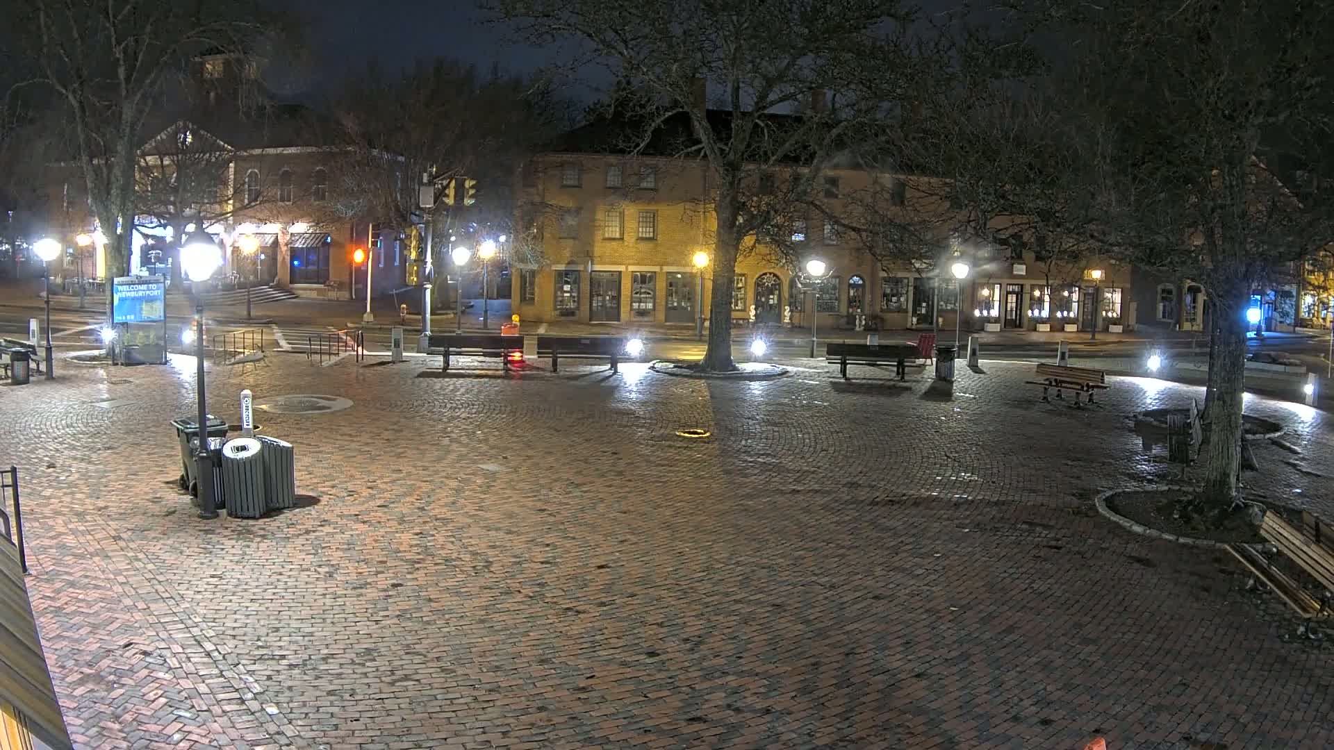 A brick town square at night, illuminated by streetlights, with several benches and trees, and a few buildings visible in the background.