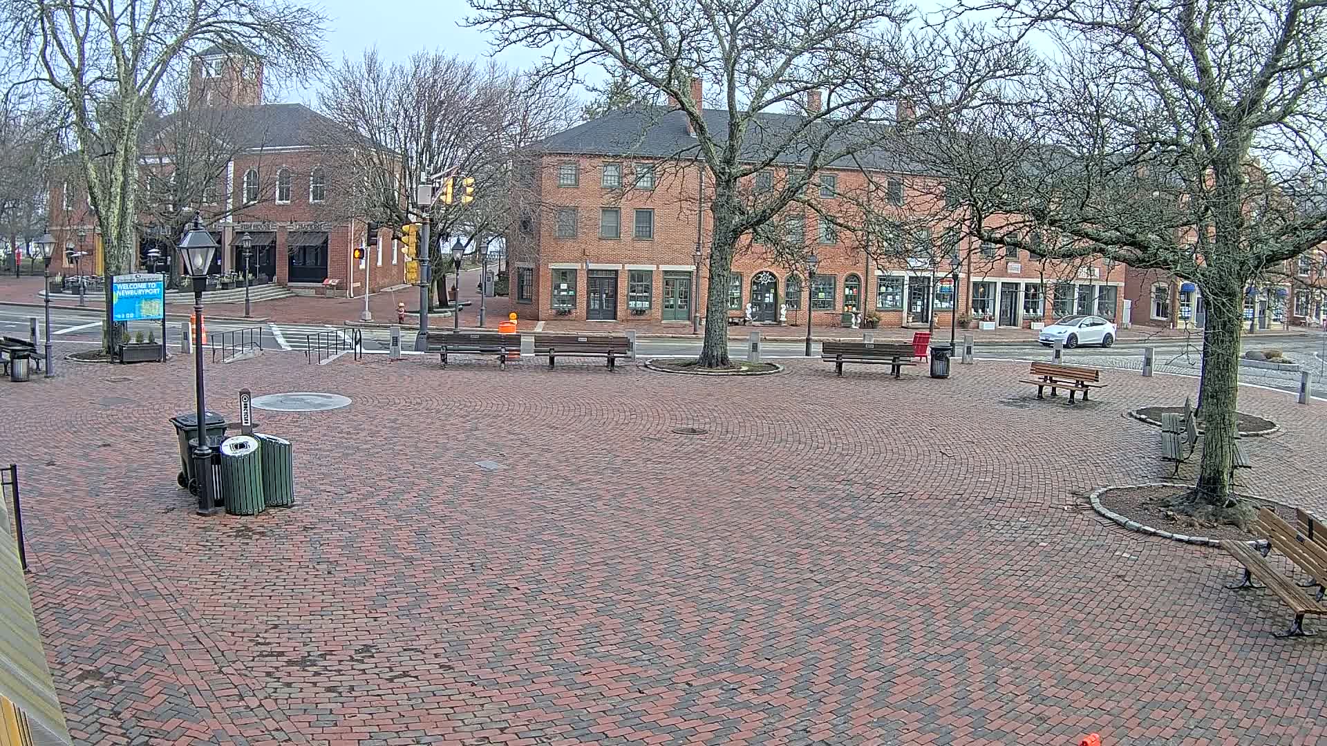A brick town square with benches and bare trees under an overcast sky, featuring brick buildings and a single car in the background.