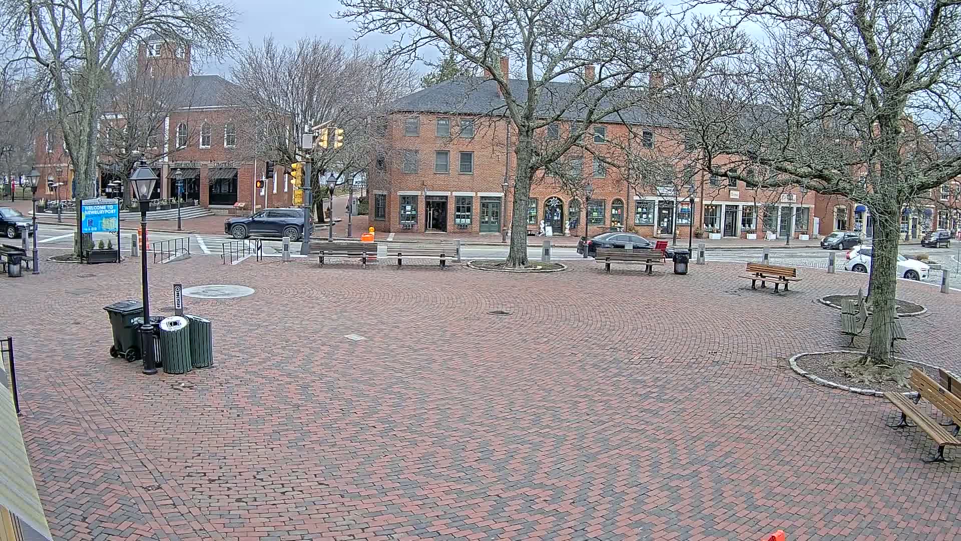 A brick plaza with several benches and trees surrounds brick buildings under a cloudy sky.